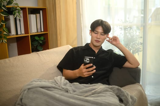 Young man listening to music in earphones and using mobile phone on sofa stay at home.