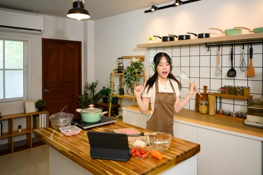Joyful young woman listening to music through headphones and dancing in the kitchen.