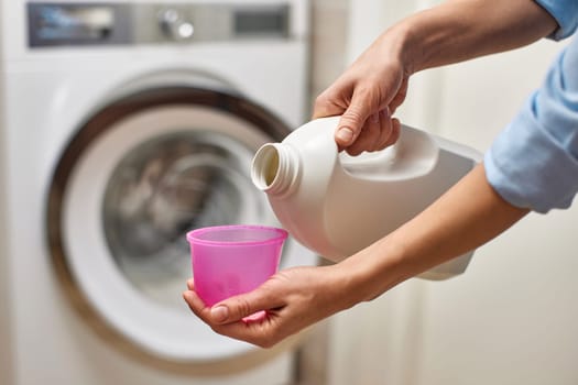Woman pours liquid transparent laundry detergent or conditioner into plastic cap against washing machine.