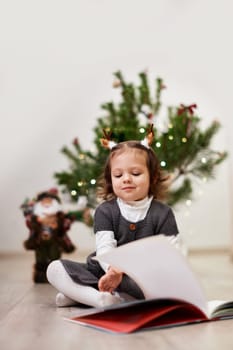 Cute little child girl reads a book near the Christmas tree at home