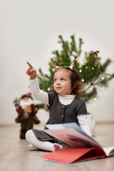 Cute little child girl reads a book near the Christmas tree at home