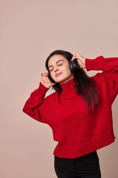 Happy woman wearing red sweater and headphones enjoying listening music on pink background.