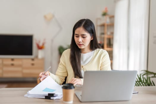 A focused young woman reviewing documents and making notes while working on her laptop in a cozy home office, emphasizing a productive atmosphere.