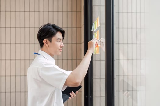 A young Asian male office worker is focused on organizing colorful sticky notes on a glass wall in a contemporary workspace, emphasizing planning.