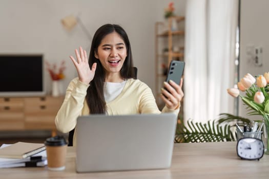 A cheerful young woman waving while having a video call in her cozy home office, exuding positivity and engagement in her workspace.