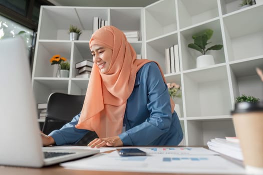 A smiling Muslim woman in a hijab organizing financial documents while working at her desk with a laptop in a bright office setting.
