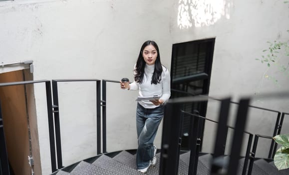 A young Asian woman is walking up a spiral staircase with a coffee cup and documents, balancing her work responsibilities and daily routine.