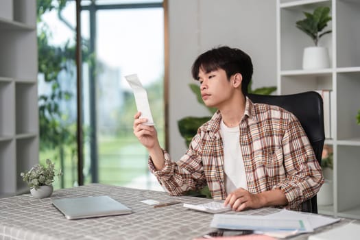 A young man examines a receipt with a focused expression while managing finances in a contemporary workspace.