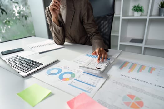 A business professional calculates finances using a calculator while analyzing reports and charts in a modern office environment.