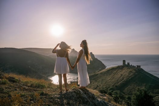 Two women are standing on a hill overlooking the ocean. They are holding hands and looking out at the water. The scene is peaceful and serene, with the sun shining brightly in the background