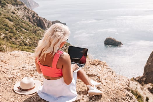 A woman is sitting on a rocky hillside with a laptop open in front of her. She is wearing a pink top and a straw hat. Concept of relaxation and leisure