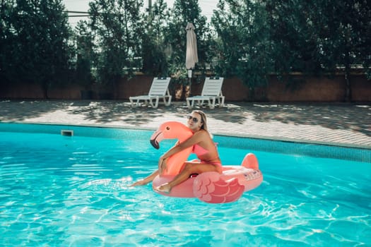 A woman is sitting on a pink flamingo float in a pool. The scene is bright and cheerful, with the woman enjoying her time in the water