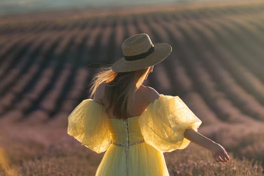 Here's a caption under 150 characters describing the image: Woman in yellow dress and straw hat, back to camera, stands in a vast lavender field at sunset.