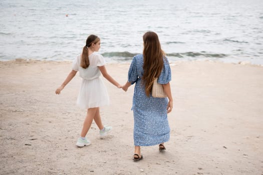 Two women, holding hands, stroll along a sandy beach towards a calm sea. One wears a white dress, the other a blue one. Summer day, rear view.