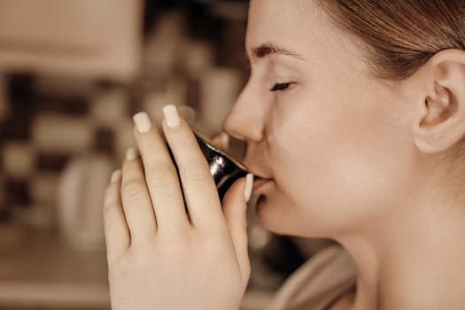 Woman with eyes closed, enjoying a warm beverage from a dark mug. Her hands gently cup the mug. Close-up profile view.