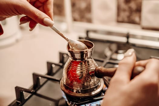 Close-up of hands adding sugar to a cezve, a traditional coffee pot, on a gas stove. The metal cezve is ornate and sits on a burner.