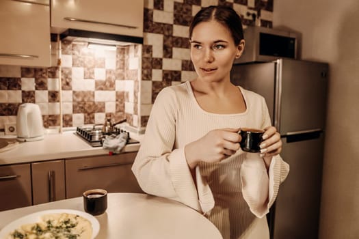 Woman in white robe stands in kitchen, holding small dark mug. A plate of food and a coffee pot are on the counter. She looks thoughtfully away.