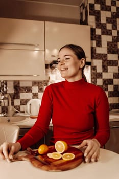 Smiling woman in a red turtleneck sits at a kitchen counter, a wooden board holding sliced oranges and a lemon before her.