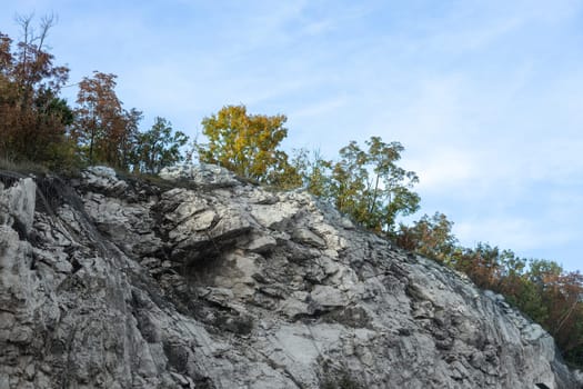 Rocky cliff with patches of green and autumn trees under a partly cloudy blue sky depicting natural erosion and geology.