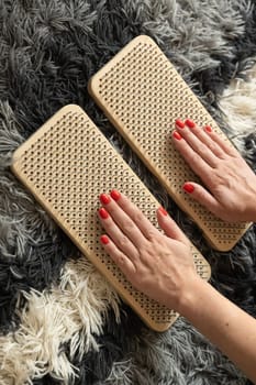 Close-up of hands with red nail polish placed on wooden acupressure massage boards, set on a textured gray and white rug. A wellness and relaxation concept.