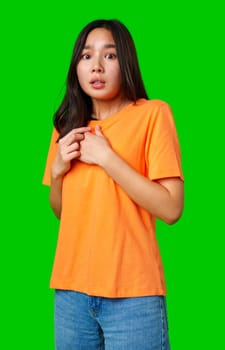 A young woman with long dark hair stands with an expression of surprise, wearing an orange shirt and casual jeans, captured against a vibrant green backdrop.