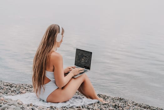 A woman is sitting on a beach with a laptop in her lap. She is wearing headphones and she is listening to music