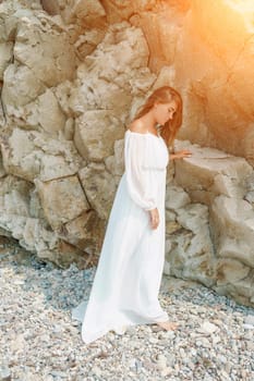 A woman in a white dress is standing on a rocky beach. Concept of serenity and tranquility, as the woman is enjoying the natural beauty of the beach
