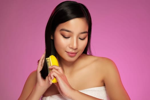 A woman with long black hair gently brushes her hair with a yellow comb, illuminated by a bold pink backdrop. She appears relaxed, showcasing an effortless beauty routine.