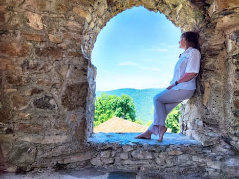 A person enjoys the tranquil mountain view from a rustic stone window on a sunny day.
