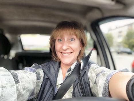 A middle-aged woman is smiling and taking a selfie while driving her car, showcasing her excitement for the trip.