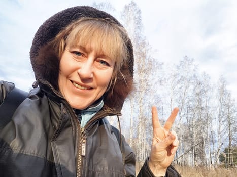 Woman enjoys nature while posing with a peace sign against a backdrop of trees and cloudy sky.