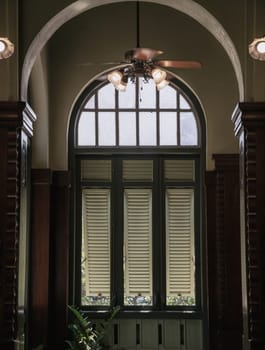 Bangkok, Thailand - Mar 02, 2025 - View of Three wooden louvered window shutters under glass frame inside of Hallway with Vintage ceiling fan and Lamps inside The main Bang Khun Phrom Palace that blends Baroque style with stucco designs. use it as your Wallpaper, Poster and Space for text, Selective focus.