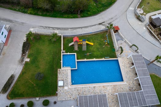 Vibrant playground equipment stands beside a clear blue pool in a lush green area under a bright sky.
