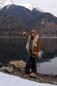 Woman in brown coat poses near a calm lake, snow-dusted rocks at her feet. Majestic snow-capped mountains form a dramatic backdrop.