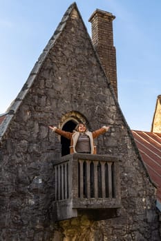 Woman joyfully spreads arms wide on a rustic wooden balcony jutting from a stone, triangular-roofed building with a brick chimney.