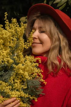 Blond woman in red hat and sweater, eyes closed, smiling, deeply inhaling the scent of a large bouquet of bright yellow mimosa flowers.