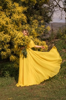 Woman in flowing yellow gown holds mimosa blossoms near a mimosa bush. Sunlight illuminates the scene. Dress billows gently.