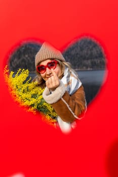 Woman in sunglasses and knit hat holds mimosa flowers, posing playfully within a heart-shaped cutout on a vibrant red background.