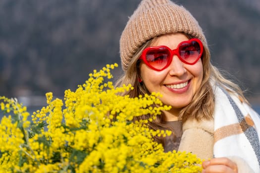 Smiling woman in a knit hat and heart-shaped sunglasses holds a large bouquet of bright yellow mimosa flowers.