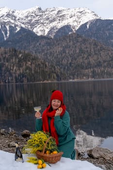 Woman enjoys a lakeside picnic amidst snowy mountains. A basket of fruit and flowers sits nearby. She holds a glass, possibly wine. Serene winter landscape.