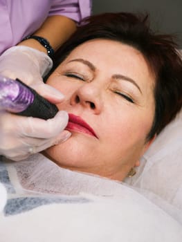 Permanent lip makeup beautiful woman over 50 years old in a beauty salon. A close-up beautician does a tattoo on his face. Vertical