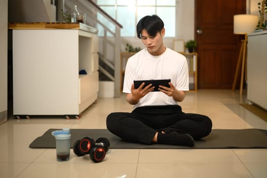 Young asian man sitting on yoga mat and using digital tablet preparing for home workout.