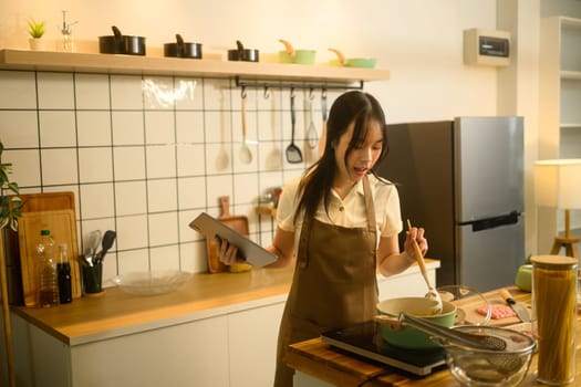Young woman following an online recipe on digital tablet while cooking spaghetti in a modern kitchen.