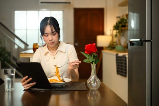 Young asian woman enjoys a plate of spaghetti and reading or browsing on digital tablet.