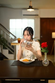Young woman drinking a glass of water and using her smartphone at the dining table.