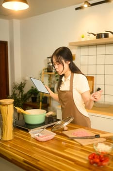 Young asian woman wearing apron holding digital tablet cooking spaghetti in a kitchen.