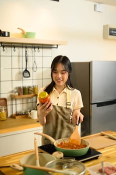Happy asian woman holding a jar of tomato sauce and stirring a pan of spaghetti sauce.