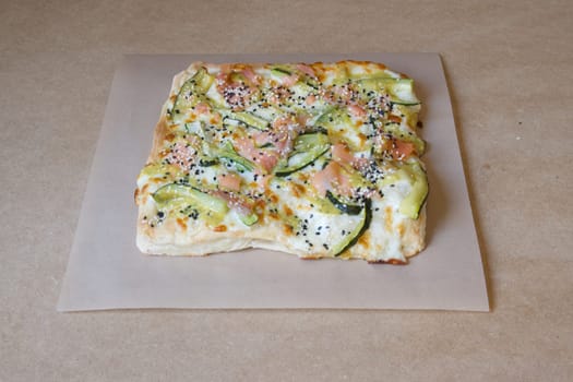 A pizza with vegetables on it is sitting on a brown table. The pizza is cut into squares and has a variety of toppings including cucumbers, tomatoes, and cheese