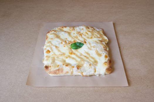 A cheese pizza with a basil leaf on top. The pizza is sitting on a brown table. The pizza is a square shape and is covered in cheese