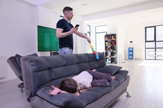 In a casual living room, a caring father uses his smartphone while cleaning around a relaxed child on the sofa